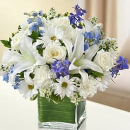 White lilies, roses, and daisies with blue flowers in a square glass vase