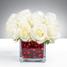 Compact arrangement of white roses in a square glass vase filled with red berries.