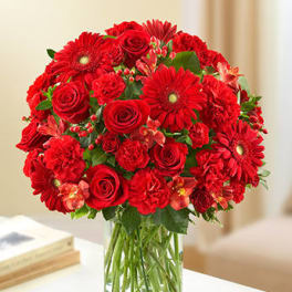 Round arrangement of red roses, gerbera daisies, and carnations in a clear glass vase