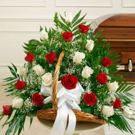 Red and white rose basket arrangement with baby's breath and fanned greenery beside a wooden casket
