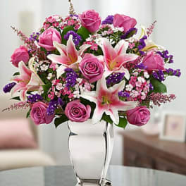 Pink roses and white lilies with purple accents arranged in a clear glass vase