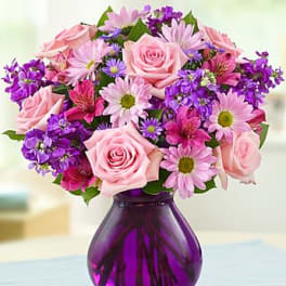 Pink roses and mixed purple and pink flowers arranged in a round purple glass vase