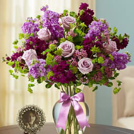 Lavender roses and purple flowers in a clear glass vase with a pink ribbon bow on a table