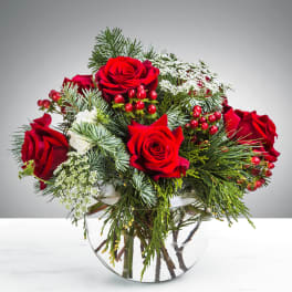 Round arrangement of red roses, white filler flowers, and evergreens in a clear glass bowl vase