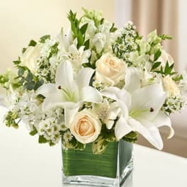White lilies, cream roses, and small white flowers arranged in a square glass vase