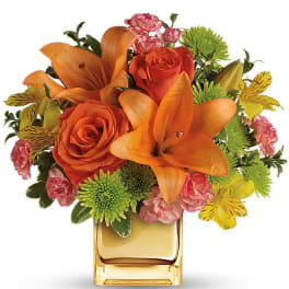 Compact arrangement of orange lilies and roses with pink and yellow accents in a gold cube vase