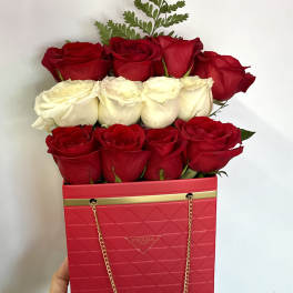 Red and white roses arranged in a pink Prada-style box with a gold chain handle.