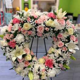 Heart-shaped standing wreath of white, pink, and cream flowers with roses, lilies, and carnations on a black easel.
