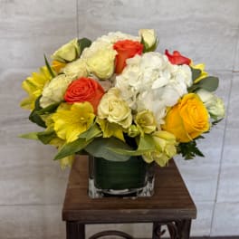 Compact arrangement of yellow, white, and orange flowers in a clear glass cube vase.
