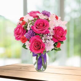 Pink roses and mixed flowers in a clear glass vase with a purple ribbon