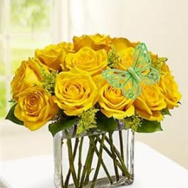 Low arrangement of yellow roses in a clear rectangular vase with a decorative green butterfly