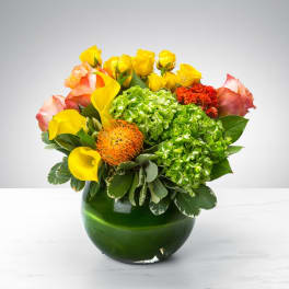 Compact arrangement of yellow roses, calla lilies and mixed blooms in a round green glass vase.