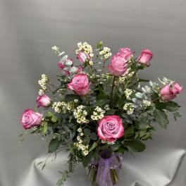 Tall arrangement of pink roses and small white blossoms in a clear glass vase with a purple ribbon
