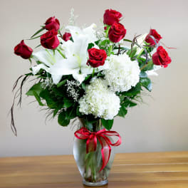 Arrangement of red roses, white lilies, and white hydrangeas in a clear glass vase with a red ribbon