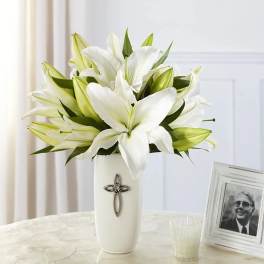 White lilies arranged in a tall white vase with a cross emblem