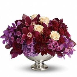Low centerpiece of cream roses, burgundy orchids, and purple blooms in a silver pedestal bowl