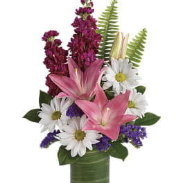 Mixed arrangement of pink lilies, white daisies, and purple flowers in a leaf-lined glass vase