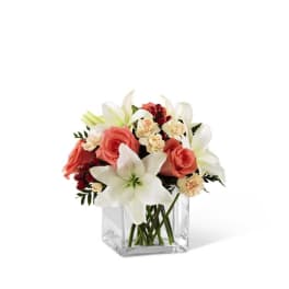 Low arrangement of white lilies, peach roses, and cream carnations in a clear glass cube vase