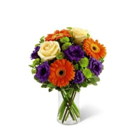 Vase arrangement of cream roses, orange gerberas, purple blooms and green button flowers in a clear glass cylinder.