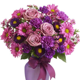 Purple and pink bouquet of roses, carnations, and daisies in a lavender vase with a matching ribbon bow