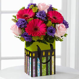 Bouquet of pink gerbera daisies, purple flowers, and pale pink roses in a decorative gift bag
