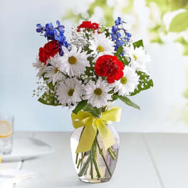 Bouquet of white daisies, red carnations, and blue flowers in a glass vase with a yellow ribbon