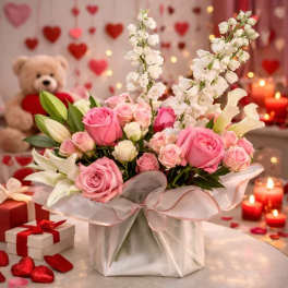 Pink roses and white lilies in a wrapped gift box with a teddy bear and candles behind
