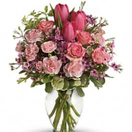 Pink tulips, roses, and carnations arranged with small purple blooms in a clear glass vase