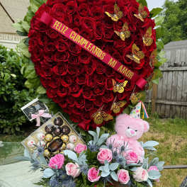 Heart-shaped red rose arrangement with pink roses, teddy bear, and chocolates