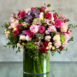Large arrangement of pink roses, peonies, orchids, and green hydrangeas in a clear glass cylinder vase