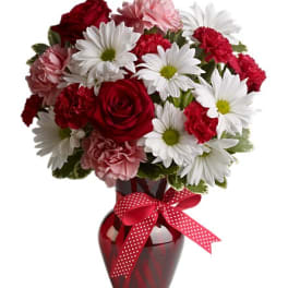 Bouquet of red roses, white daisies, and pink carnations in a red vase with a ribbon