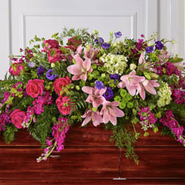 Bright pink lilies, roses, and purple blooms arranged as a casket spray on a wooden casket