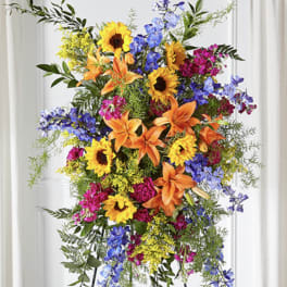 Tall standing spray with sunflowers, orange lilies, blue delphinium, and bright pink blooms on an easel