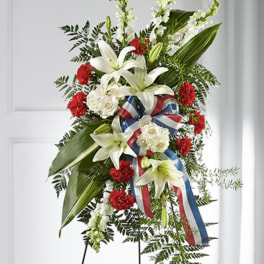Tall standing spray with white lilies, white roses, red carnations and a red, white, and blue ribbon on an easel