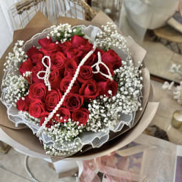 Hand-tied bouquet of red roses and white filler flowers wrapped in paper with draped pearl strands.