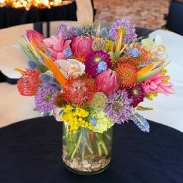 Bright mixed tropical and protea flower arrangement in a clear glass vase with stones.