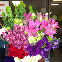 Tall mixed floral arrangement in a green vase with pink lilies, orchids, and red roses