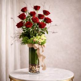 Tall arrangement of red roses and green orchids in a clear glass vase with river stones and burlap bow.