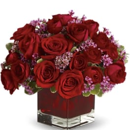 Red roses arranged in a square glass vase with small purple filler flowers