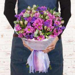 Handheld bouquet of pink daisies and purple filler flowers wrapped in lavender paper