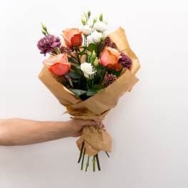 Handheld bouquet of peach, white, and purple flowers wrapped in brown paper