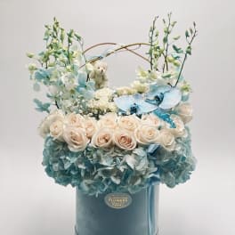 Blue and white floral arrangement in a round hatbox with a small teddy bear