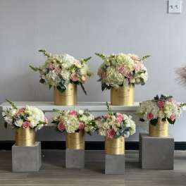 Five pastel floral arrangements in gold cylindrical boxes on gray pedestals