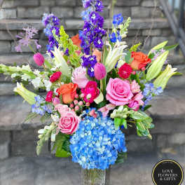 Tall mixed bouquet with pink roses, lilies, blue hydrangea, and purple delphinium in a clear vase