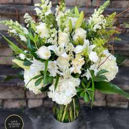 Tall arrangement of white roses, lilies, hydrangeas, and snapdragons in a clear glass vase.
