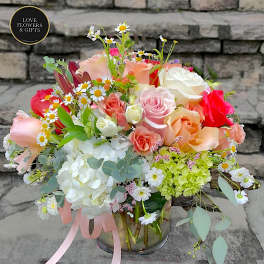 Low glass vase of mixed roses, hydrangeas, and daisies in soft pink, peach, white, and green tones