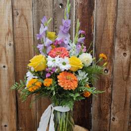 Tall mixed bouquet of orange, yellow, and purple flowers in a clear glass vase with white ribbon.