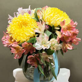 Yellow and pink flowers arranged in a glass vase with a blue ribbon
