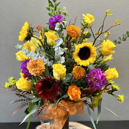 Bright mixed bouquet with sunflowers, yellow roses, and purple blooms in a glass vase