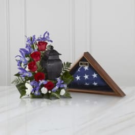 Red roses and purple flowers arranged beside a folded American flag in a wooden case.